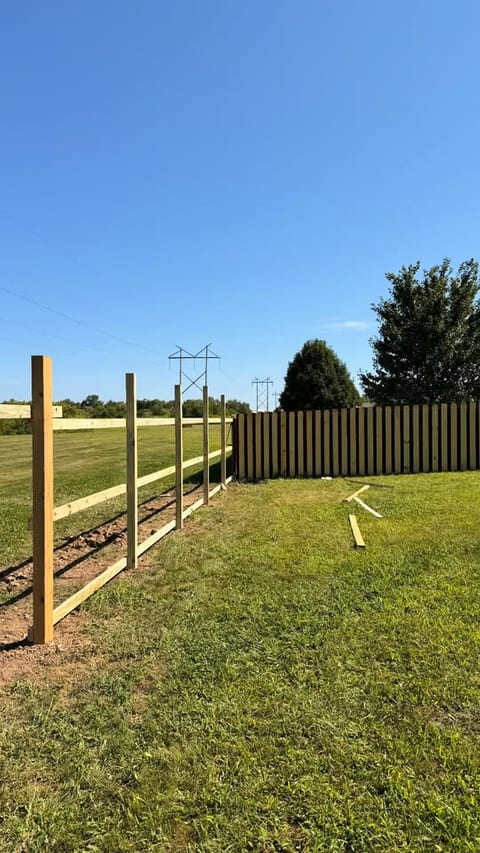 Pressure-treated fence posts and rails set in a line on a rural Indiana property, with a completed dark-stained privacy fence running behind them.